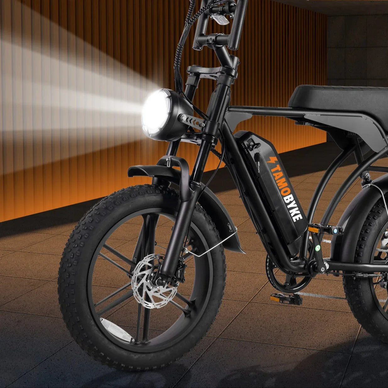 Front view of a black electric bike with fat tires and headlight on, indoors at night
