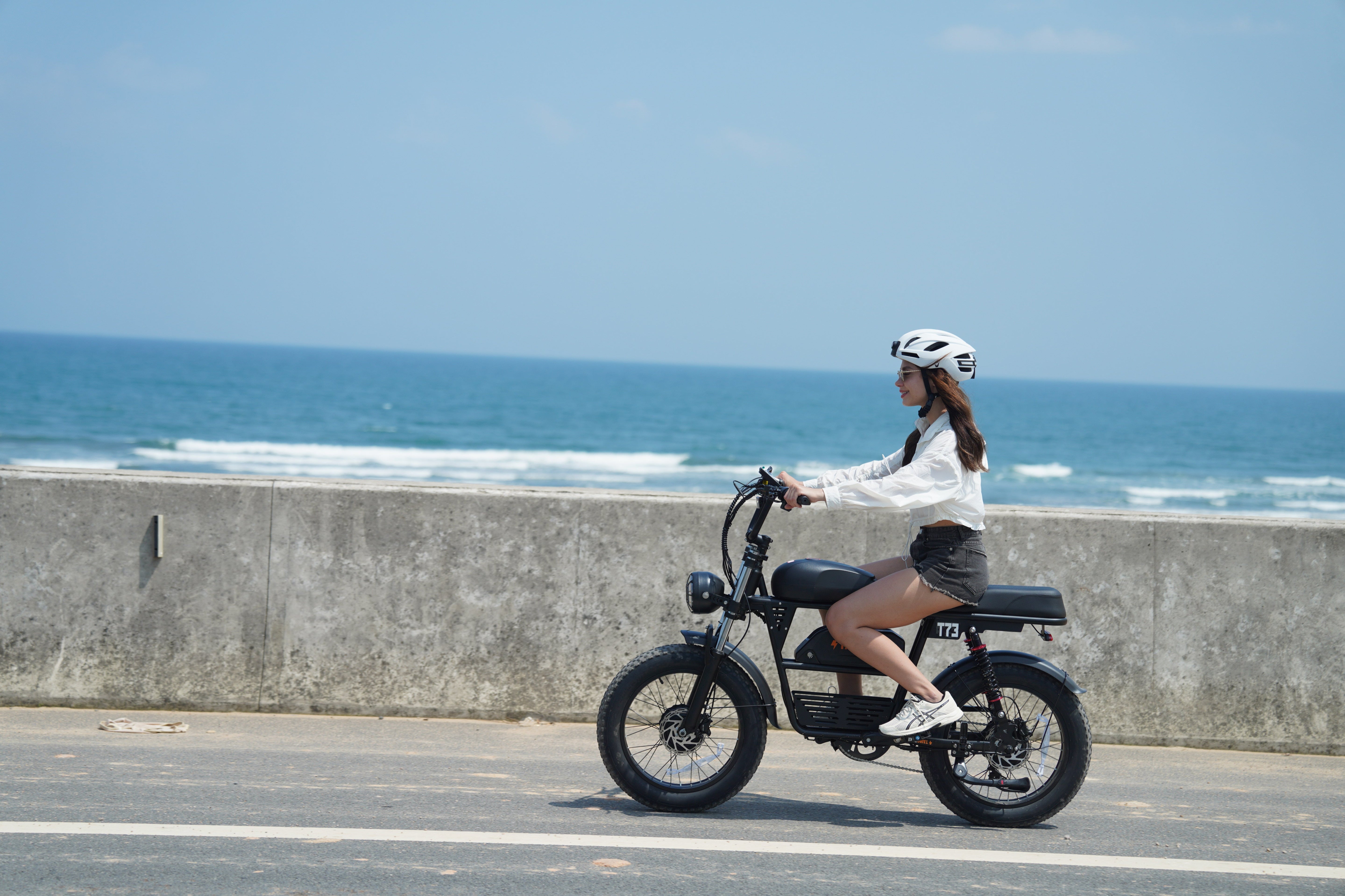 Woman riding the Tamobyke T73 Pro Max electric bike along a coastal road,highlighting its stylish design